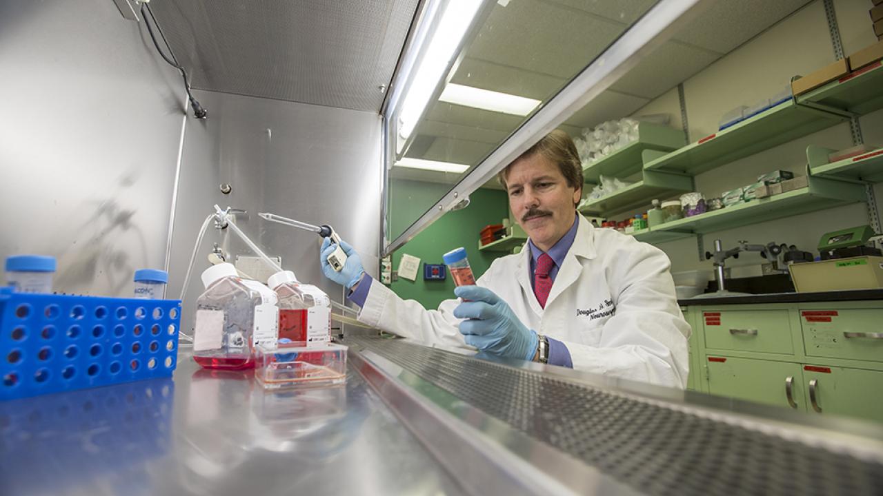 Dr. Douglas Smith wears a white lab coat and blue gloves while sitting at a lab fume hood stocked with reagents.